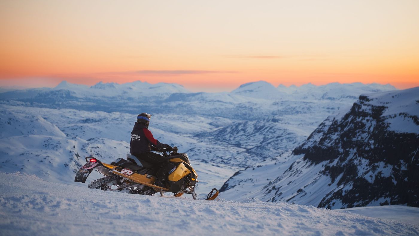 Förare på en gul Ski-Doo som blickar ut över en snötäckt fjälldal i kvällsljus