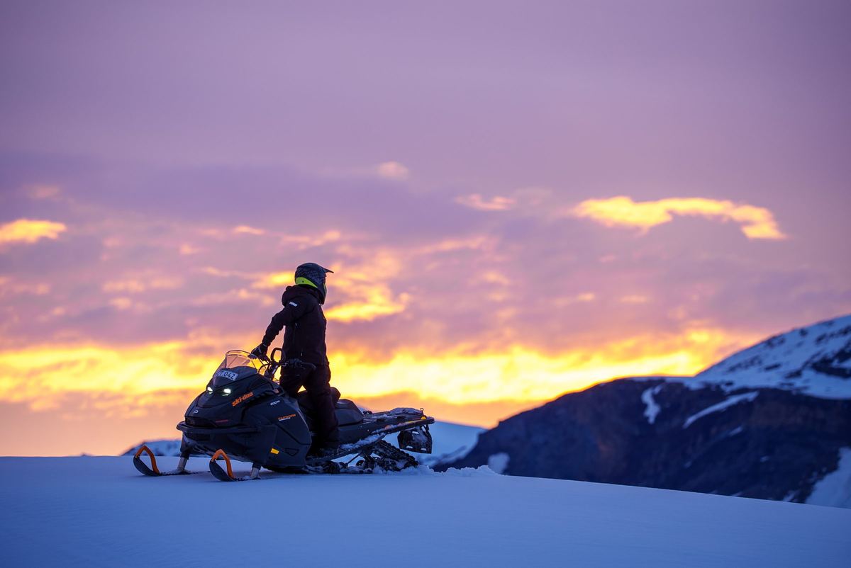 Förare som står på en svart Ski-Doo på fjället i solnedgången